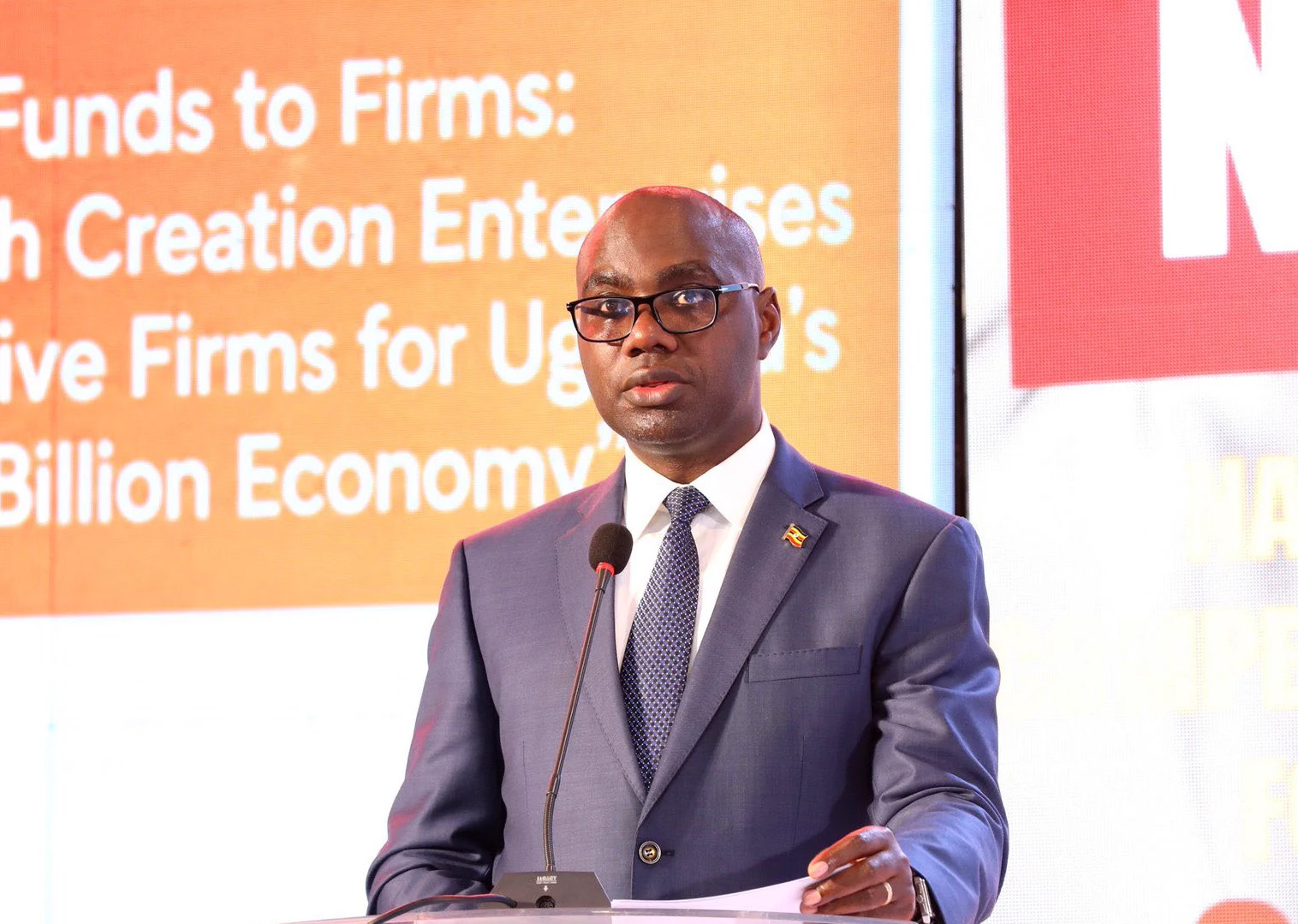 Finance Minister Henry Musasizi speaking at a lectern in front of a conference banner